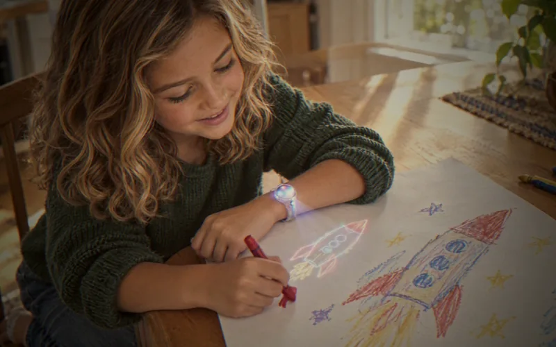 At the kitchen table, a six-year-old traces a rocket that Halo projects onto her paper, beside her own crayon drawing.